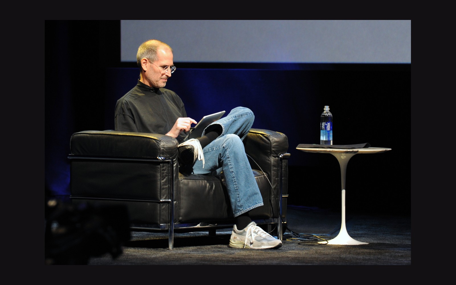 A man in a black turtleneck sits in a leather chair, using a tablet onstage